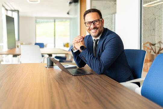 Professional man sitting at an office desk with a laptop, smiling at the camera, representing success, business expertise, and corporate leadership in a modern workspace