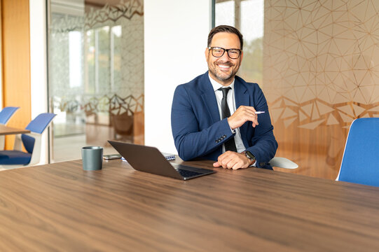Confident businessman smiling. Holding a pen. And sitting at a dark conference table with a laptop. Reflecting corporate leadership. Professional services