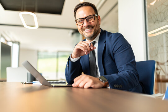 Smiling professional businessman looking directly at the viewer. Sitting confidently at a modern office desk with a laptop
