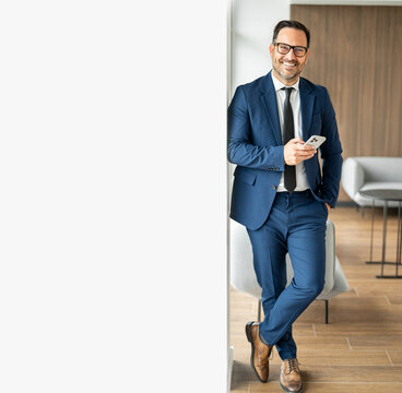 Businessman in blue suit leaning against a wall in a contemporary office setting, holding a mobile device and looking confidently at the viewer