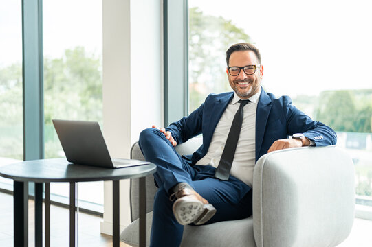 Confident business person in a suit smiling, sitting relaxed in a contemporary office setting with a laptop nearby, reflecting success, achievement, and a professional workspace