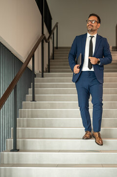 Professional businessman in a blue suit and black tie descending a modern staircase, carrying a tablet, looking confident and successful in a corporate environment