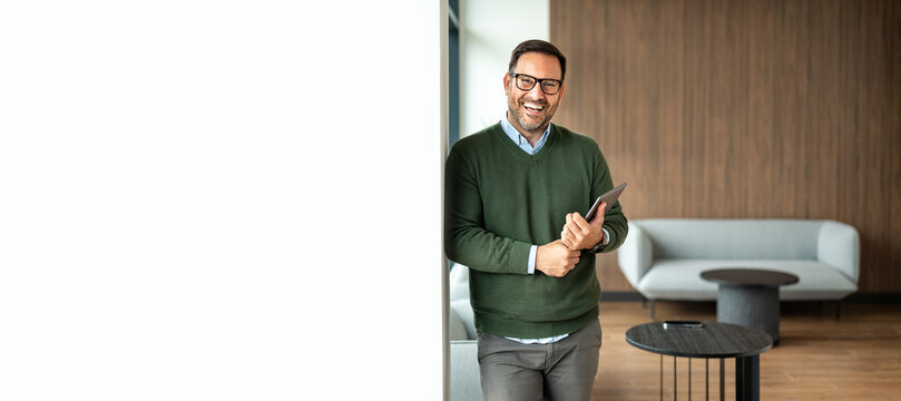 Smiling businessman holding a digital tablet in a professional and minimal office lounge area, portraying a successful and confident corporate professional utilizing technology