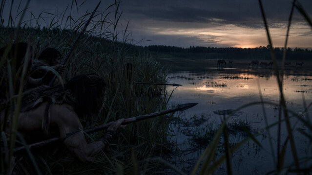 Prehistoric Hunt at Dawn: Two primitive hunters crouch low in reeds, spears poised, as they prepare to ambush prey near a tranquil body of water during the early morning light.