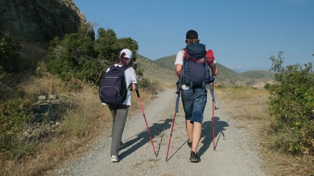 Camera follows father and teenage daughter hiking with backpacks and trekking poles on a mountain road, family hiking adventure, outdoor endurance training and hiking skills