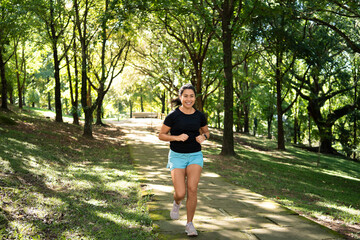 Naklejka premium Woman, running and smiling in park with fitness, wide shot or trail path outdoor. Front view, athlete or runner with smart watch, nature and cardio training in forest