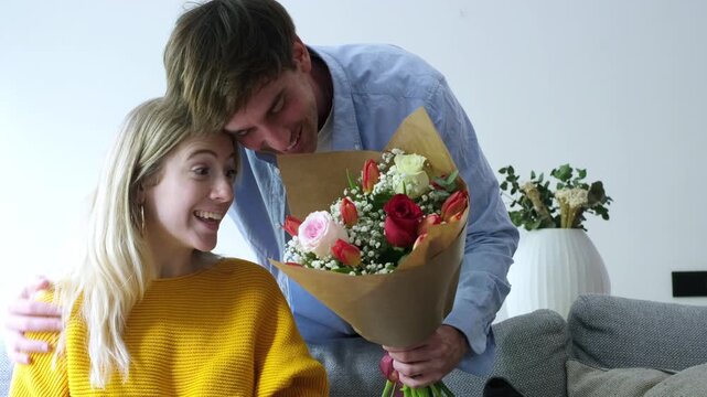 A loving boyfriend surprises his happy girlfriend with a beautiful bouquet of fresh flowers, including roses and tulips, in their home. Her face lights up with joy and excitement as she receives the r