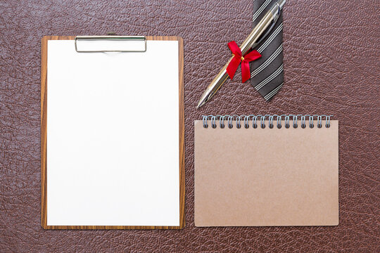 Blank white report paper on wooden clipboard and notebook with pen and necktie on brown leather background, business and education concept