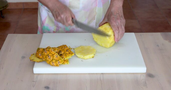 Female hands cutting a peeled fresh pineapple into round slices with a kitchen knife on a white cutting board.