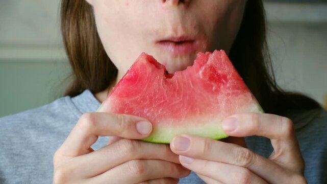 Young hungry woman biting fresh slice of watermelon, enjoying its sweet, refreshing taste. Close up mouth showing enjoyment of healthy summer fruit snack at home. Woman eating juicy watermelon slice