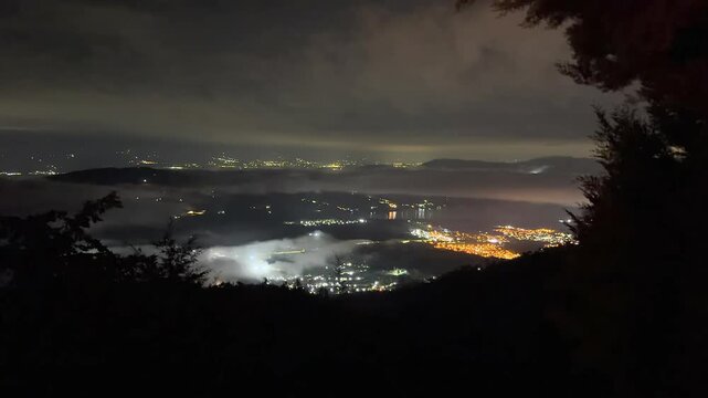 night aerial view of illuminated city and valley under fog and clouds