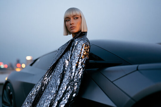 Blonde Frau in silbernem Metallic-Outfit lehnt an futuristischem Auto