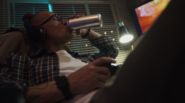 Caucasian young adult man wearing headphones playing video game with controller and drinking from aluminum can at night. Clip ideal for ads on digital addiction or gaming lifestyle