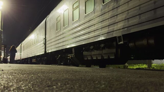A passenger train waits at the station platform at night. A woman walks near the illuminated cars of Russian Railways. This mysterious and peaceful scene feels adventurous.