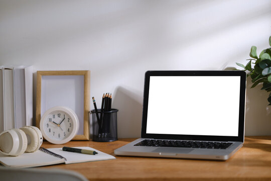 Modern home office workspace with blank laptop screen, notebook, headphones, clock and stationery arranged on a wooden desk with natural light.