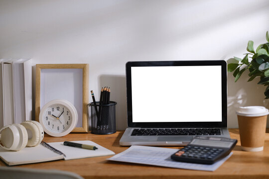 Modern home office workspace with laptop mockup screen, calculator, notebook, headphones, coffee cup and stationery on wooden desk.