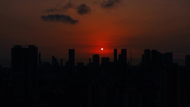 Mumbai skyline during sunset, with the sun setting behind a dense cluster of high-rise buildings and the Arabian Sea visible on the horizon. Warm orange sky creates a dramatic urban silhouette.