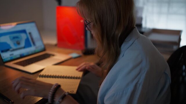 Teenage girl wearing eyeglasses flipping through a notebook at a desk beside an active laptop in the evening. Education, studying, homework and academic learning concept.