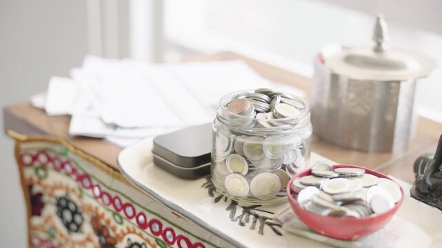 Glass jar and red bowl overflowing with mexican peso coins on a decorated desk at home