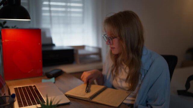 Dedicated female student working late, thoughtfully chewing on a pen before getting an idea and smiling while taking notes in a workbook beside a laptop.