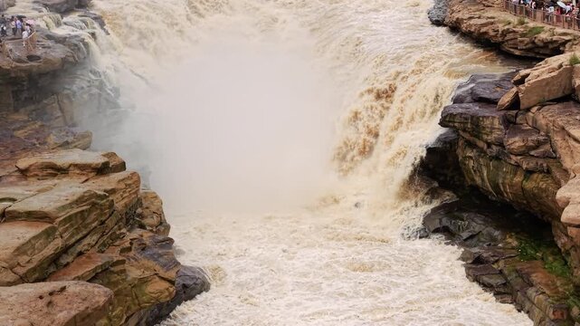 Powerful muddy river plunging into a rocky gorge. A turbulent frenzy of splashing water and mist, showcasing nature's raw energy and fluid dynamics in a strict static composition.