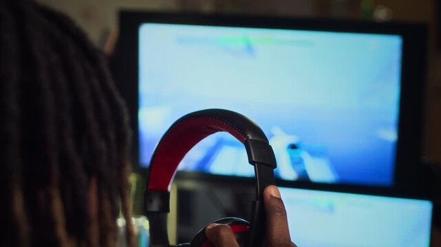African American young adult man putting on headphones while playing shooter video game on computer screen, rear view close up. Footage perfect for gaming addiction or esports campaigns