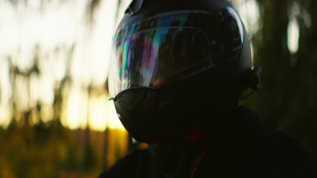 Motorcyclist wearing reflective visor helmet rides through tranquil forest at sunset, silhouetted against warm golden light and tall trees in peaceful scenery