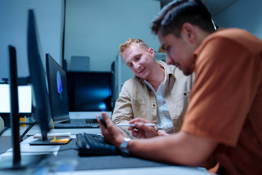 Young male IT software developer smiling while his colleague uses a pen to point at a digital tablet screen during a programming system development workflow in a modern office setup.