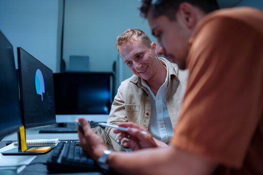 Happy young male IT developers smiling while pointing at a screen, collaborating on programming code and website system design in a modern computer laboratory workspace.