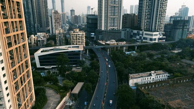 Cinematic aerial drone footage showing Mumbai's dense urban landscape, with modern high-rise buildings, older residential structures, an elevated metro line, busy roads and ongoing construction.