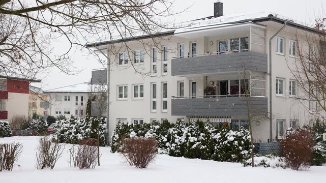 Snowy residential yard in Germany shows modern apartment buildings, balconies, hedges and bare trees during winter snowfall. Quiet urban neighborhood reflects cold weather, housing and seasonal life