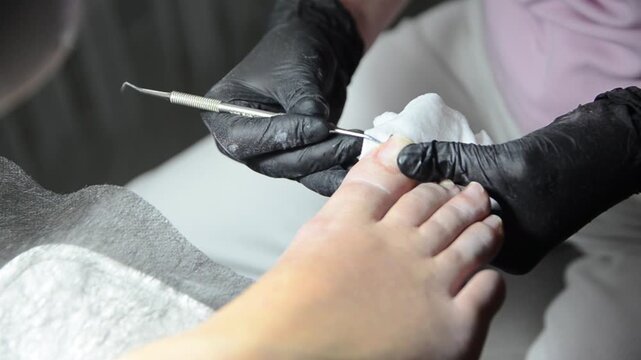 Pedicurist carefully cleans under the toenail with a metal curette during a professional pedicure procedure. Close-up foot and nail care in a beauty salon.