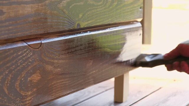 Hand in glove painting wooden box with brown oil stain brush. Worker applying protective varnish on pine timber planks surface, DIY woodwork. Wood staining process. Backyard Woodworking, Maintenance.