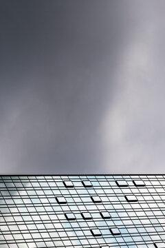 Dark clouds fill the sky above a low viewpoint as windows recede in a pattern of strict geometry along the facade, turning modern urban architecture into an upward view.