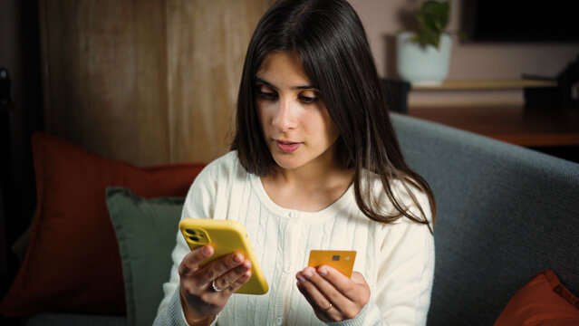 Young Latina woman shopping online during Cyber Monday from her sofa, using her smartphone and payment card to complete a purchase at home.