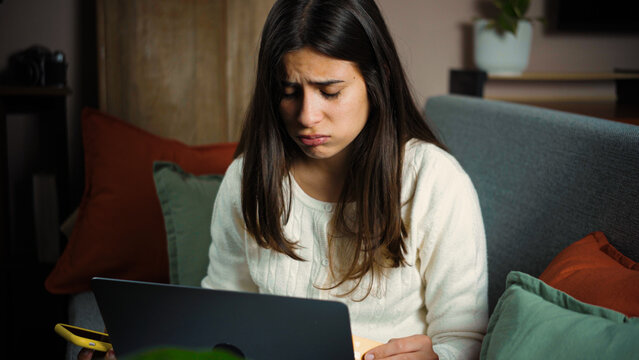 Young Latina woman sitting on her sofa with her laptop, looking very frustrated and overwhelmed by a problem on her computer.