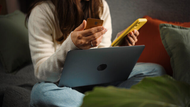 Anonymous young woman sitting on her sofa and shopping online with her payment card during Cyber Monday, browsing deals and completing a purchase from home.