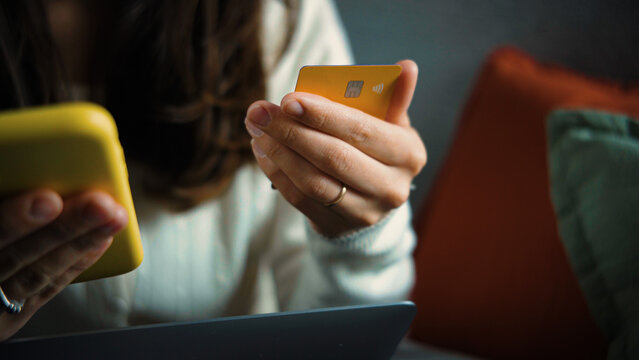 Close-up of an anonymous woman's hand holding a credit card while sitting on a sofa and preparing to make an online purchase from home.