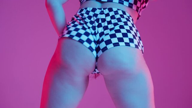 Young woman dancing twerk in checkerboard shorts, vibrant studio with colored lights. Modern dance performance, confidence, rhythm and expressive stage presence. Twerking