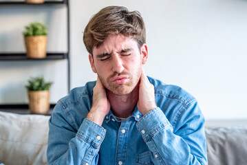 Fototapeta premium Young man with neck pain holding his neck, wincing in discomfort on a couch at home