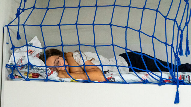 Person sleeps on a loft bed with a blue net above in a simple room during the daytime