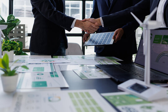 Business people closing a deal, shaking hands over a table with esg reports, a solar panel, and a wind turbine model