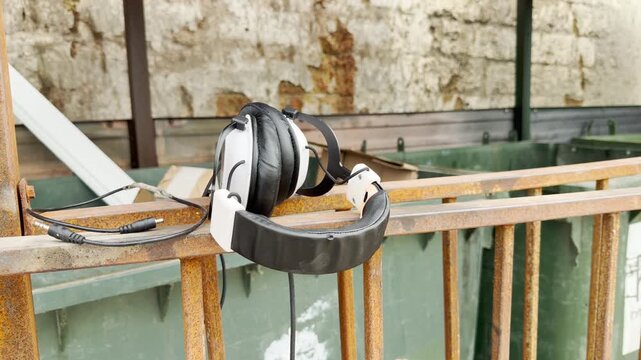 White headphones hang on a rusty metal railing near trash bins outdoors. The discarded headset rests silently in daylight. The lonely scene evokes a sad, nostalgic feel.