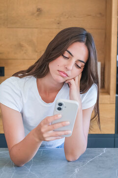 Young woman in white t-shirt looking bored and scrolling smartphone at kitchen counter, feeling distracted and disengaged while using social media and texting, casual lifestyle portrait
