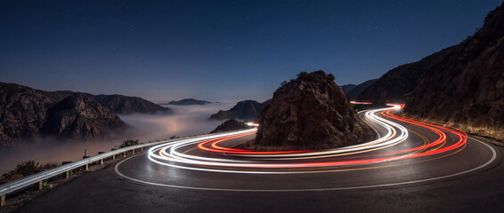 Light trails on a winding mountain road captured with long exposure night photography, featuring vibrant red car streaks around a rock bend under starry skies for travel banners © itlada