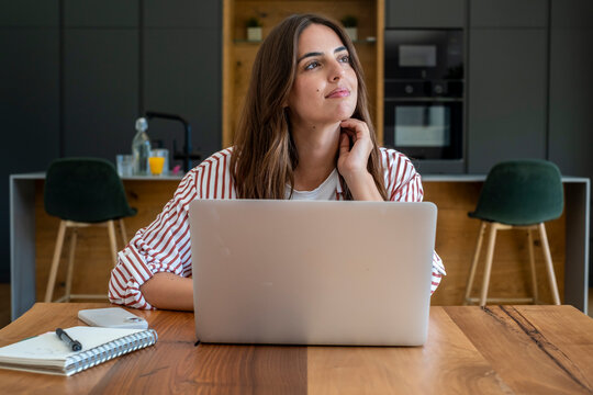 Young woman working on a laptop in a modern kitchen, looking thoughtful while studying or doing remote work at a wooden table