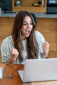 Excited young woman celebrating success while working on a laptop at home, cheering with clenched fists and joyful expression, remote work achievement and online win