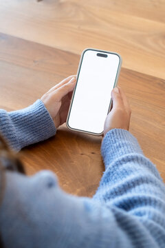 Person holding smartphone with blank white screen over wooden table, wearing blue sweater &mdash; close-up of hands using mobile device for browsing, messaging, app mockup and everyday tech use