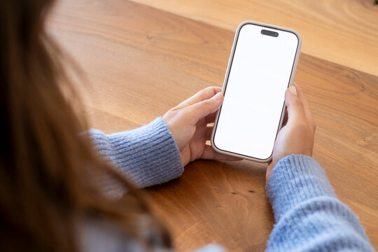 Person holding smartphone with blank white screen over wooden table, hands in blue sweater ready to use mobile phone