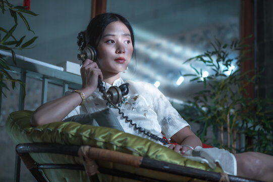 Pretty young Chinese woman wearing white silk qipao and classic necklace talking on vintage phone, posing gracefully in retro style interior with warm nostalgic ambient lighting